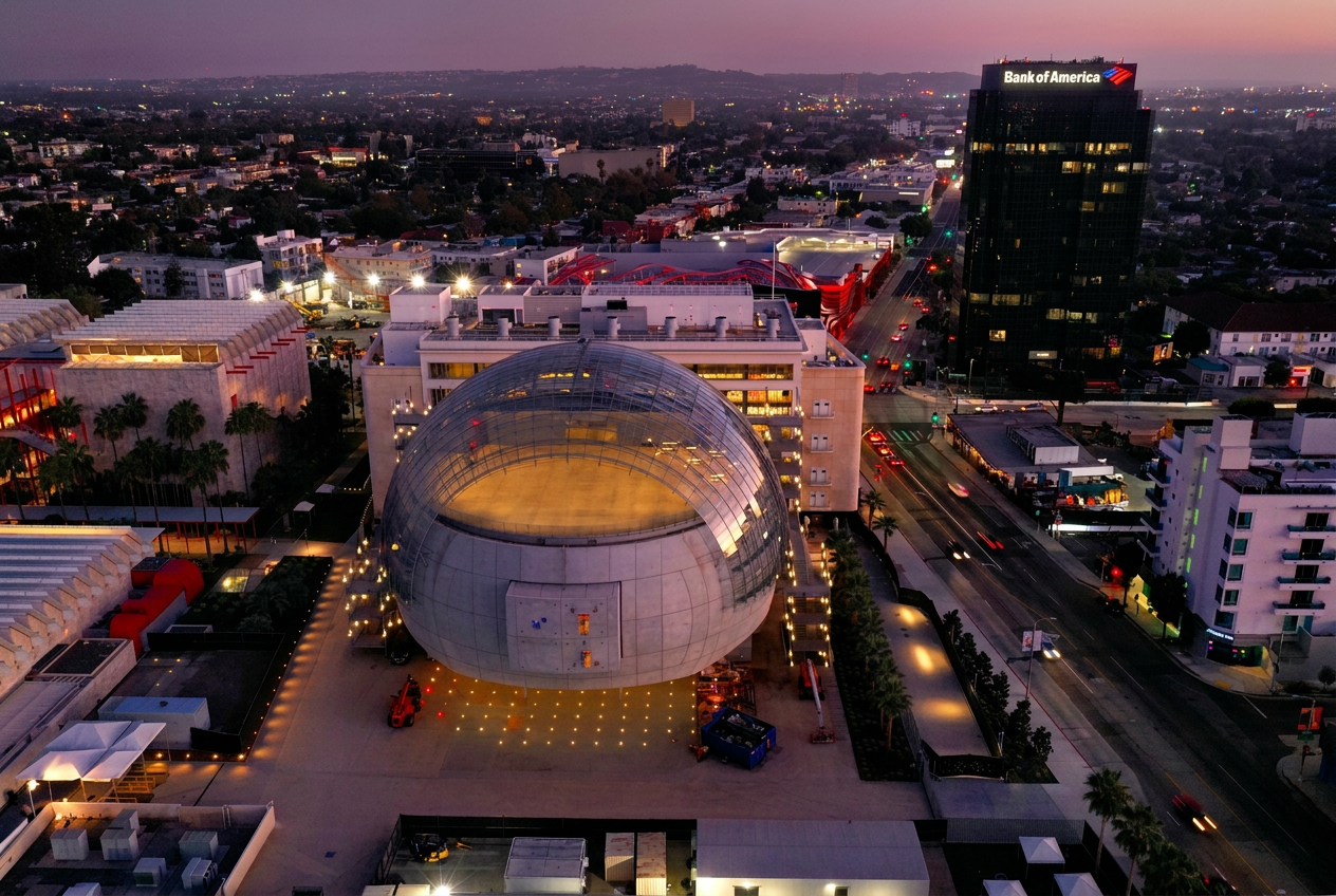 Aerial view at dusk of the Bank of America Tower at 6100 Wilshire Boulevard, with the Academy Museum of Motion Pictures sphere and Petersen Automotive Museum in the foreground along Museum Row